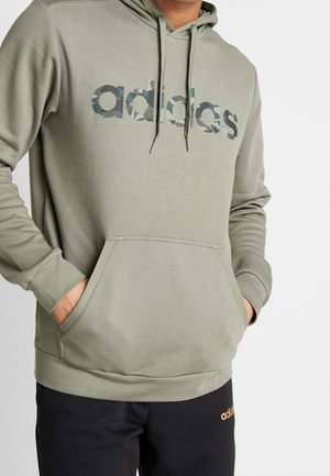 Mężczyzna w oliwkowo-zielonej bluzie z kapturem Adidas z logo w kamuflażowym wzorze oraz czarnych spodniach Adidas z pomarańczowym logo, ręce w kieszeni bluzy.