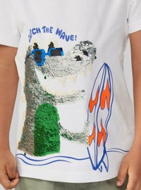 Weißes T-Shirt mit einem Pailletten-Krokodil, das eine blaue Sonnenbrille trägt, sowie einem bunten Surfbrett und dem Text "Catch the Wave!" in Blau.