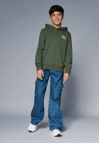 Grüne Kapuzenjacke mit einer vorderen Tasche und gelbem Logo, kombiniert mit blauen weiten Cargo-Jeans mit seitlichen Taschen und weißen Sneakern.