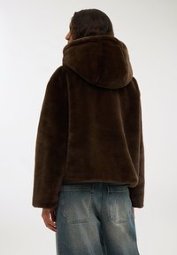 Giacca in pelliccia ecologica marrone con un ampio cappuccio, spalle basse e una consistenza morbida. Abbinata a jeans in denim azzurri chiari con tasche posteriori.