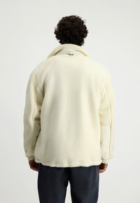 Kurtka polarowa w kolorze kremowym, z wysokim kołnierzem, elastycznymi mankietami oraz miękkim, teksturowanym materiałem o luźnym kroju.