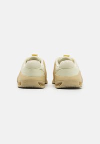 Adidași galbeni deschiși și bej, cu un superior texturat și talpă din cauciuc, având un dop la spate și un branding subtil.