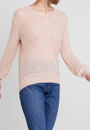 Frau trägt einen hellrosa gestrickten V-Ausschnitt-Pullover mit langen Ärmeln und blaue Jeans, steht vor einem schlichten Hintergrund.