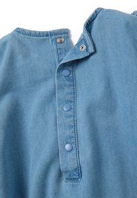 Tessuto di denim blu con colletto arrotondato, caratterizzato da cinque bottoni in plastica blu sul davanti e dettagli di cucitura visibili lungo i bordi.
