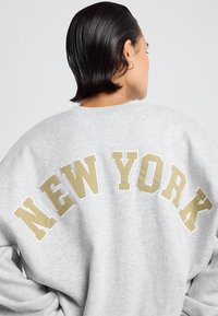 Persona con cabello corto y oscuro que lleva un sudadero gris claro con "NEW YORK" en letras doradas grandes en la parte de atrás.