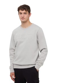 Felpa grigia chiara con stampa testurizzata "REALITY" sul petto, collo rotondo e polsini e orlo a costine. Abbinata a pantaloni neri.