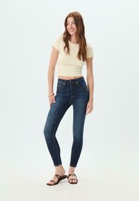 Tmavě modré skinny džíny s jemným prošitím, vysokým pasem a předními kapsami; spárované s přiléhavým krémovým crop topem a plochými hnědými sandály.