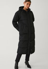 Zwarte gewatteerde jas met capuchon, met een lange snit, quilted textuur en drukknopsluiting. Twee zijzakken zichtbaar.