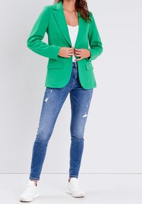Woman wearing green blazer, white top, distressed skinny jeans, and white sneakers, standing against a plain light background.