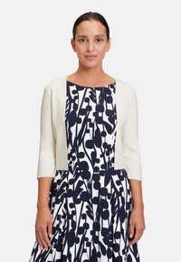 White cropped jacket with three-quarter sleeves over a navy and white floral-patterned dress. Smooth texture, modern design, and fitted shape.