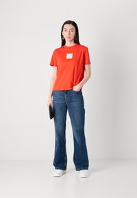 Rode katoenen t-shirt met korte mouwen en een blauw grafisch logo aan de voorkant, gecombineerd met vloeiende donkerblauwe jeans en witte sneakers.