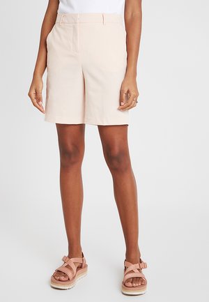 Pantalones cortos ajustados de color rosa claro con corte recto, parte delantera plana y bolsillos laterales. Llevados con sandalias rosas con tiras y suelas blancas.