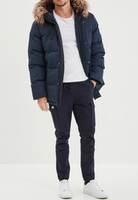 Navyblauer Pufferjacke mit Kapuze mit Fellbesatz, Reißverschluss vorne und Taschen, kombiniert mit dunklen Hosen und weißen Turnschuhen. Darunter ein schlichtes weißes T-Shirt.