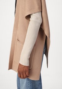 Persona che indossa un gilet in maglia beige senza maniche con tasca frontale sopra una maglietta a maniche lunghe bianca a coste e jeans blu, con la mano rilassata lungo il fianco.
