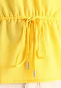 Top amarillo texturizado con cintura ajustable mediante cordón con puntas de plata, creando un efecto de fruncido en la línea de la cintura.
