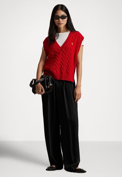 Rdeč pleten top brez rokavov z V-izrezom in vzorcem kitke, nošen prek bele majice, kombiniran s črnimi širokimi hlačami in črno torbico.