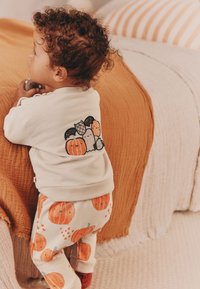 Sweat-shirt beige clair avec un motif de citrouille dans le dos, assorti à un pantalon crème orné de motifs orange de citrouilles. Textures confortables.