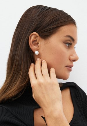 Woman with long brown hair wearing a black top, touching a large white pearl earring on her right ear, against a white background.