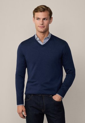Navy v-neck sweater with long sleeves, made of a textured fabric. Worn over a light blue and white striped shirt at the collar. Dark jeans.