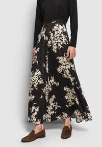 Frau in einem schwarzen Maxikleid mit großen weißen Blumenmustern, einem schwarzen Langarmoberteil und braunen Loafern im Leopardenmuster.