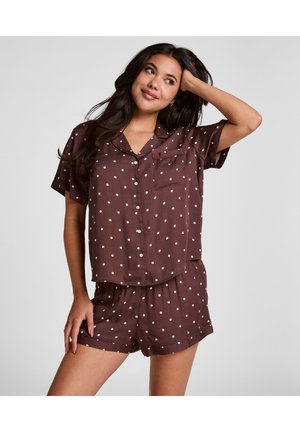 Femme aux longs cheveux foncés portant une chemise marron à pois à manches courtes et un short assorti, posant avec une main sur la tête.