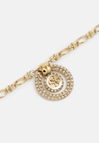 Pulsera de tono dorado con un colgante que presenta dos diseños circulares concéntricos, adornados con cristales transparentes, y un logo de marca en el centro.