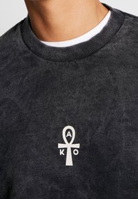 Felpa grigia scura con collo tondo e tessuto testurizzato. Presenta un simbolo dell'ankh bianco con le lettere "A" e "K" sotto di esso.