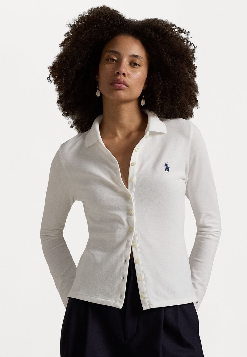 White long-sleeve polo shirt with a collar, featuring a button placket, subtle logo, and soft texture. Worn with dark pants.