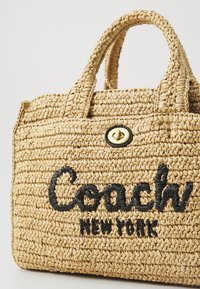 Tkaná slamenná kabelka s dvoma uchami, s čiernou vyšitou nálepkou "Coach NEW YORK" a detailom zlatého kovania.