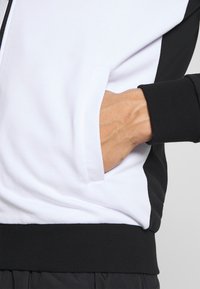 Giacca con cappuccio caratterizzata da un corpo bianco con dettagli neri, polsini a coste e una tasca laterale. Il materiale sembra morbido con una superficie liscia.