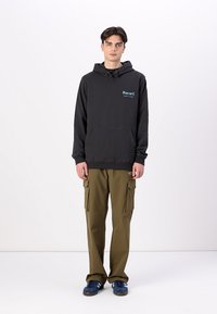 Schwarzer Hoodie mit Fronttasche und einem mehrfarbigen Logo. Kombiniert mit olivgrünen Cargo-Hosen und blauen Sneakers. Einfaches, lässiges Design.
