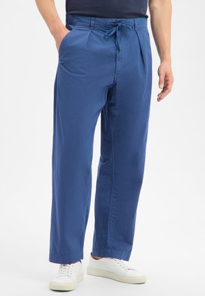 Mann trägt locker sitzende blaue Hose mit Kordelzug und Taschen sowie weiße Sneakers, steht vor einem einfarbigen Hintergrund.