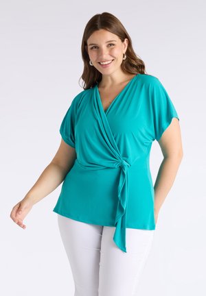 Top avvolgente color turchese in tessuto leggero con maniche corte e dettaglio con nodo laterale, abbinato a pantaloni bianchi. Texture liscia e vestibilità comoda.