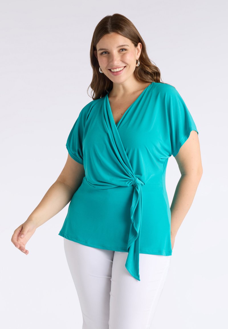 Top avvolgente color turchese in tessuto leggero con maniche corte e dettaglio con nodo laterale, abbinato a pantaloni bianchi. Texture liscia e vestibilità comoda.