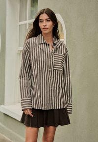 Camicia a righe marrone e bianca con bottoni, taschino sul petto, maniche lunghe con polsini, abbinata a una gonna scura plissettata.