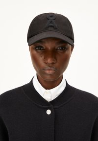 Schwarze Mütze mit einem gestickten Buchstaben "A" aus dunklem Stoff, kombiniert mit einer schwarzen Strickjacke und einem weißen Hemd mit Kragen. Insgesamt glatte Texturen.