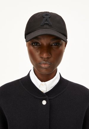 Casquette noire avec une lettre "A" brodée en tissu sombre, associée à une veste en tricot noire et une chemise à col blanc. Textures lisses dans l'ensemble.