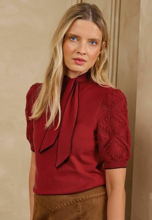 Vrouw met lang blond haar draagt een rood gestructureerd top met korte mouwen en een strikdetail in de nek, en een bruine broek met hoge taille.
