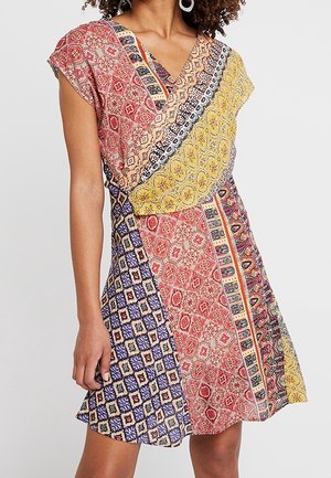 Vrouw draagt een korte mouw patchworkjurk met V-hals en geometrische en bloemmotieven in rood, geel, blauw en beige.