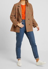 Blazer à carreaux marron à double boutonnage, coupe oversize, associé à un haut en maille orange et un jean en denim bleu, complété par des baskets blanches.