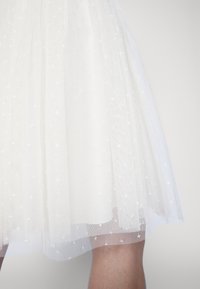 Jupe en tulle blanc avec petits motifs d’étoiles portée au-dessus des genoux, montrant une vue partielle des jambes nues sur un fond clair uni.