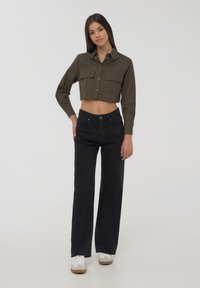 Camicia tagliata color verde oliva con due tasche sul petto, abbinata a jeans larghi neri a vita alta e sneakers bianche con dettagli marroni.