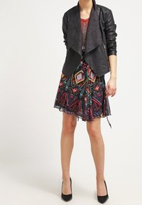 Giacca nera in pelle sintetica con fronte drappeggiato. Sotto, un vestito a fantasia colorata con design geometrici, lungo fino al ginocchio e con un orlo fluente.