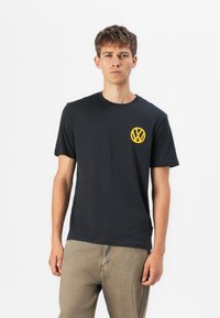 Sort kortærmet t-shirt lavet af bomuld med et gult Volkswagen-logo på venstre bryst. Løs pasform med rund halsåbning.
