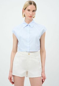 Camicetta corta a maniche corte di colore azzurro chiaro, con bottoni e strisce verticali. Abbinata a pantaloni corti di jeans bianchi con orlo sfrangiato.