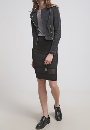 Mujer con chaqueta corta de cuero negra, camiseta gris, falda negra con panel transparente y zapatos negros con cordones, de pie sobre un fondo liso.