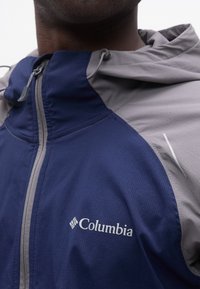 Κοντινό πλάνο ενός ατόμου που φοράει ένα μπλε και γκρι μπουφάν Columbia με φερμουάρ και ρυθμιζόμενο κουκούλα.