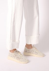 Lace-Sneaker in Creme mit einem strukturierten Muster und beiger Gummisohle, kombiniert mit weißen, umgeschlagenen Hosen.