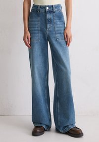 Persoon in hoge taille, wijde blauwe jeans met grote voorzakken en donkerbruine platform schoenen, staande tegen een effen muur.
