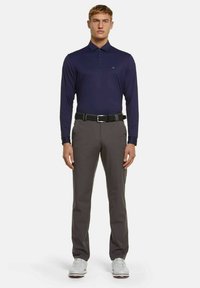 Dunkelblaues Langarm-Polohemd mit Kragen, kombiniert mit grauen Hosen und einem schwarzen Gürtel. Füße in grauen Sportschuhen mit weißen Akzenten.